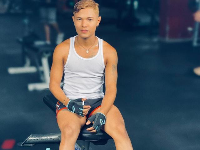 HotTonny sits on a gym bench, wearing a white tank top, red shorts, black gloves, and a necklace. He has short blonde hair and a muscular build.