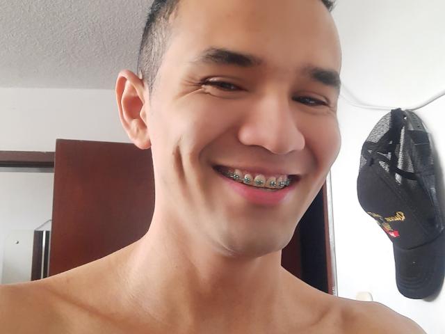 AndrewHotX has a cute smile with metal braces, short dark hair, and is shirtless, showing a smooth, tan chest. Black cap with a logo hangs in the background.