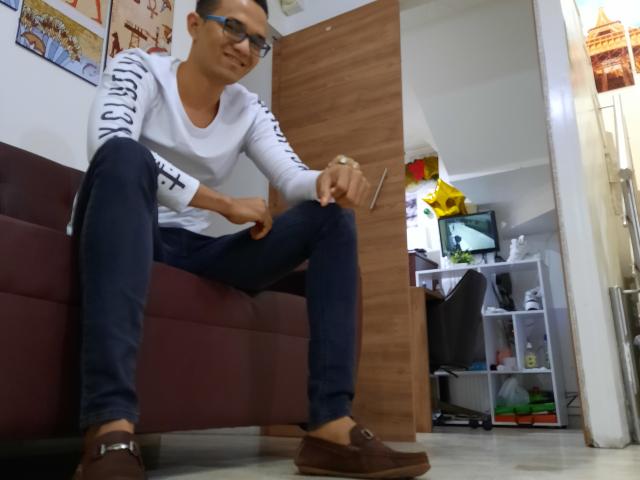 AndrewHotX, wearing glasses and a white long-sleeve shirt with black stripes, sits on a maroon couch, adjusting brown loafers, with blue jeans and a relaxed smile.
