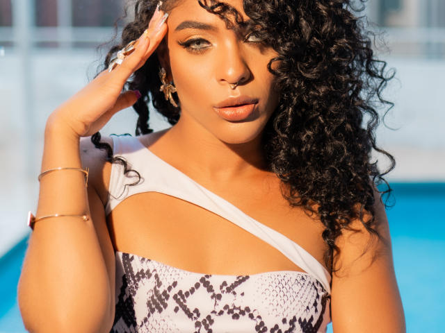 VioletHotX with curly black hair, tan skin, and snake-print dress, touching her face, showing off her nose ring and gold bracelet, with blue background.
