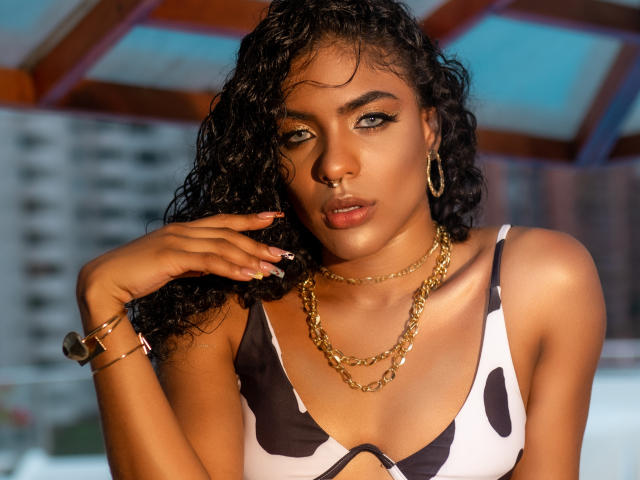 VioletHotX with curly black hair, gold hoop earrings, gold chain necklaces, and a black-and-white cutout swimsuit, touching her face, showing her dark eyebrows and shiny lips.