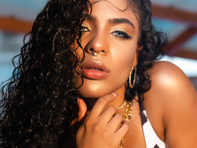 VioletHotX has dark curly hair, golden eyeshadow, and a nose ring. She's wearing a gold chain and hoop earrings, with her hand near her face, looking sexy.