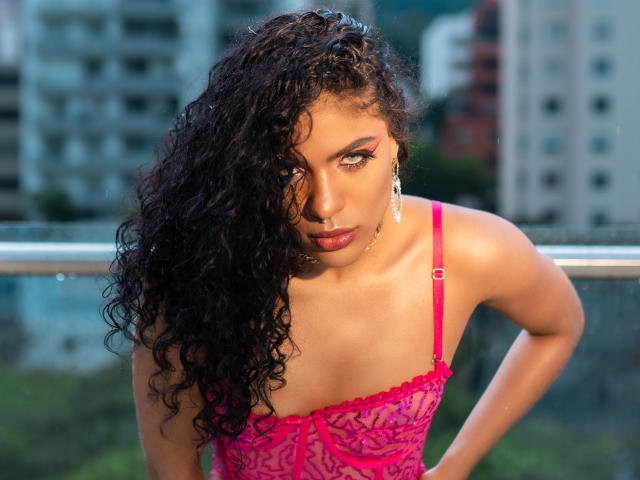 VioletHotX with curly black hair, pink lace lingerie, and serious eyes, leaning forward, looking at the camera, wearing dangling earrings, with a sultry, sexy expression.