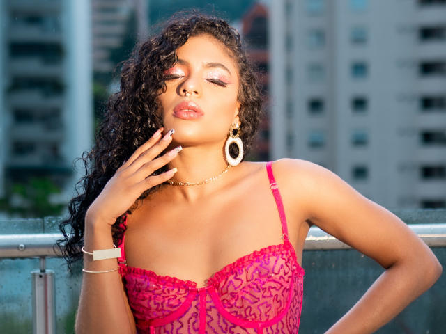 VioletHotX with curly black hair, pink lace bra, and gold hoop earrings, touching her cheek, wearing pink lipstick and gold bracelet, standing against a metal railing.