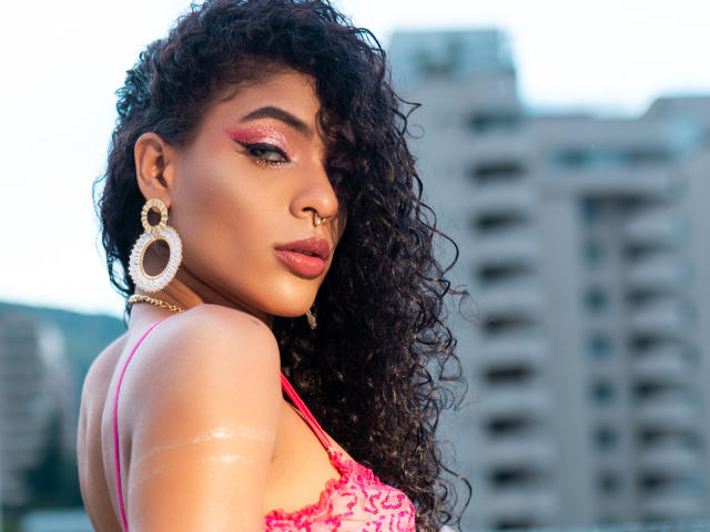 VioletHotX with curly black hair, pink makeup, gold earrings, nose ring, and pink lace top, showing off shiny tattoos and a sexy, confident look. Urban background blurred.