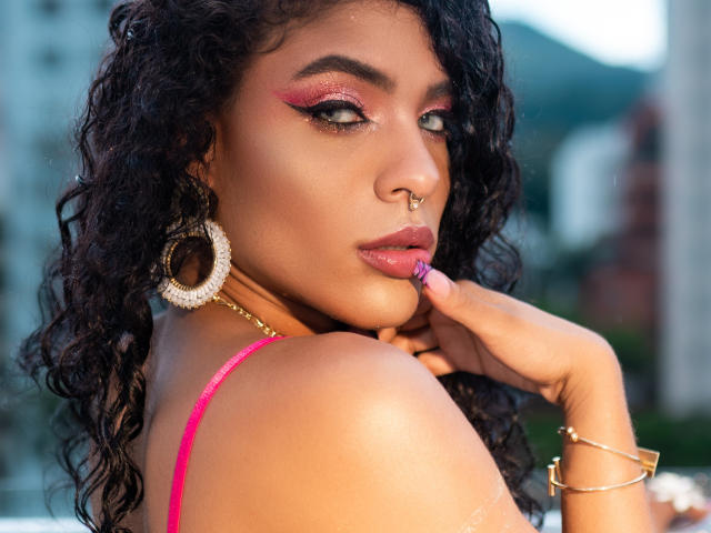 VioletHotX with curly black hair, pink eyeliner, and nose ring, touching her pink-lipped, glossy lips with her finger, wearing a pink top and gold hoop earrings.