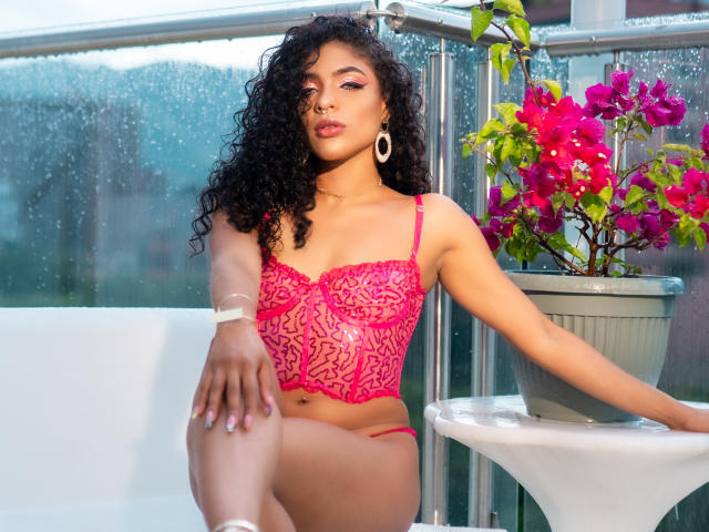 VioletHotX with curly black hair, wearing pink lace lingerie, sitting on white chair, leg up, arm on gray pot with pink flowers, white earrings, looking at camera.