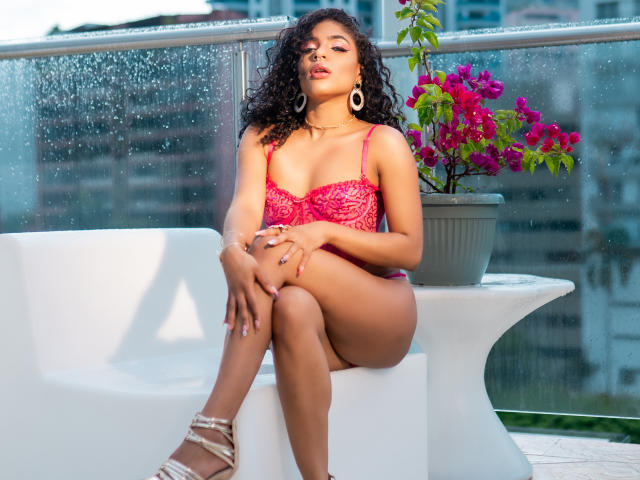 VioletHotX sits on a white chair, wearing a red lace lingerie set, showing her tits, with curly hair, large hoop earrings, and white strappy heels, under a potted plant.