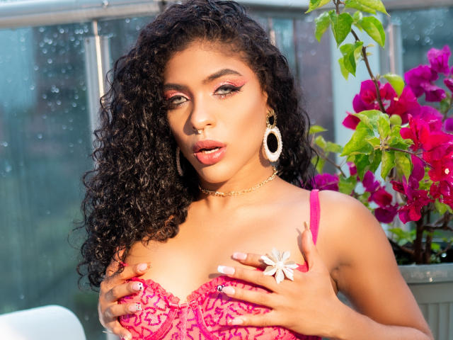 VioletHotX with curly black hair, pink lace bra, covering her tits with hands, wearing white flower pin, gold earrings, and pink eyeliner, in sunny garden with purple bougainvillea.