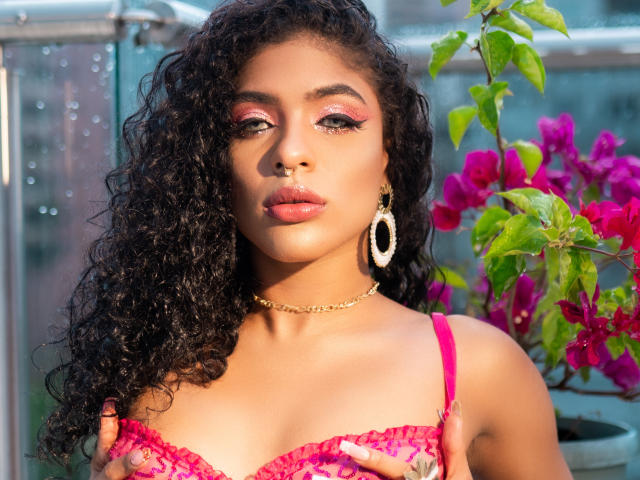 VioletHotX with curly black hair, pink eyelids, gold hoop earrings, gold chain necklace, pink lace bra, touching her small tits, standing near bright pink bougainvillea.
