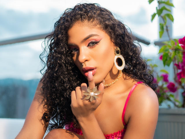 VioletHotX with curly black hair, pink makeup, and red bra, finger in mouth, gold earrings, and necklace, showing off her tits and cute nose ring. Bright sunlight.
