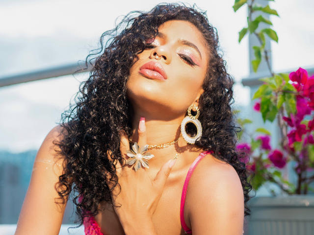 VioletHotX with curly black hair, eyes closed, pink lipstick, gold earrings, and pink top, hand near tits, sunlit, floral necklace, pink flowers in background.