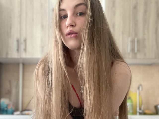 Doris hat langes, glattes blondes Haar, volle, rosafarbene Lippen und grüne Augen, trägt einen roten Träger und blickt leicht nach unten. Sie befindet sich in einer Küche und hat ein süßes, attraktives Gesicht..