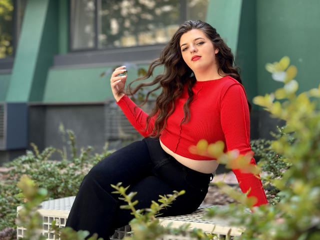 PamDares con lunghi capelli castani ondulati, top rosso, pantaloni neri, seduta su una panchina di metallo, con una mano tra i capelli, sfondo di un edificio verde, sguardo sicuro, rossetto rosso, sexy..