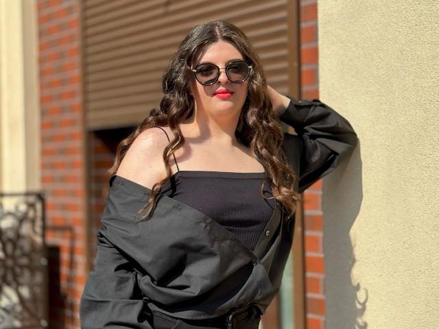 PamDares with wavy brown hair, black off-shoulder top, and black jacket, wearing dark sunglasses and red lipstick, leaning against a yellow wall, hand in hair.