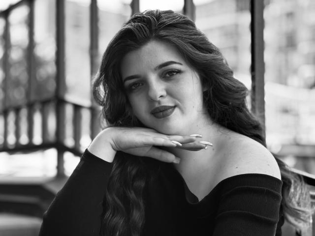 Fotografía en blanco y negro de PamDares con cabello oscuro y ondulado, una blusa negra con un hombro descubierto, apoyando el mentón en la mano, sonriendo suavemente, mostrando sutilmente sus glúteos y senos..