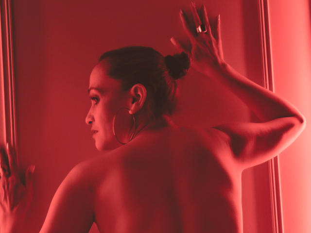 AgathaStone with her back to the camera, topless, red-lit, hands against a wall, hoop earrings, bun, ring, and a confident, sexy pose. Her ass is partially visible.