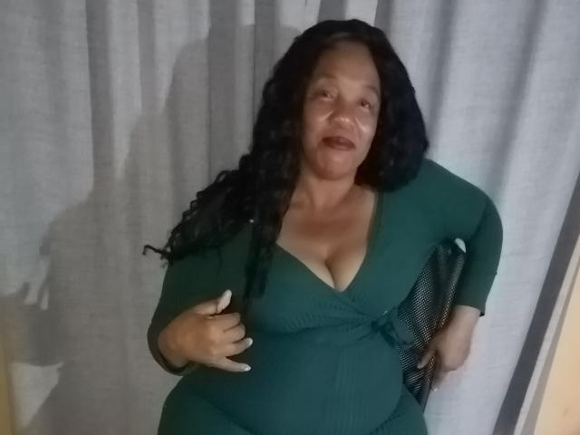 BigMarry has curly black hair, a green dress with a deep V, showing her tits, and a pregnant belly, standing against a gray curtain. Her hand is on her belly.