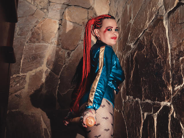MorganaCrowly with red braids, wearing a shiny blue jacket, fishnet pants, and holding a butt plug, standing against a stone wall, looking back with red makeup.