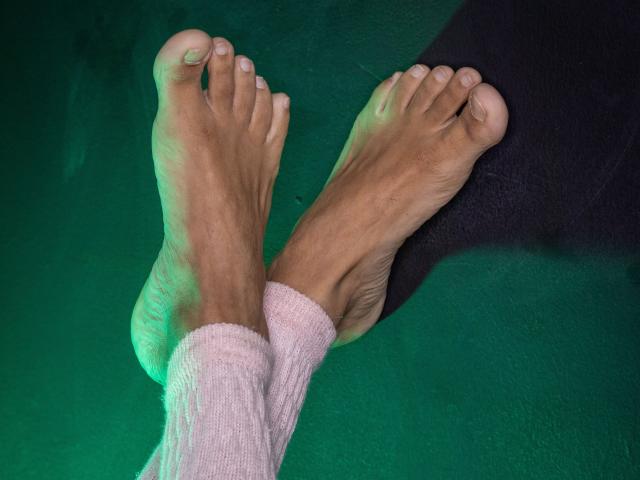 Los pies descalzos de ToroHot, cubiertos con un calcetín rosa, acanalado y de punto que llega hasta las pantorrillas, cruzados en los tobillos, mostrando ligeramente los dedos sucios contra un fondo iluminado en verde..