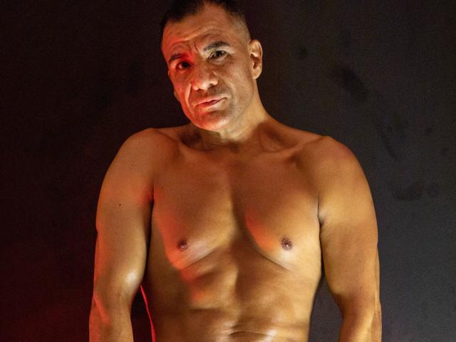 ToroHot es un hombre musculoso, sin camisa, con un tono de piel bronceado, cabello corto y oscuro, y pezones pequeños y duros. Está mirando a la cámara con una sonrisa leve y segura..