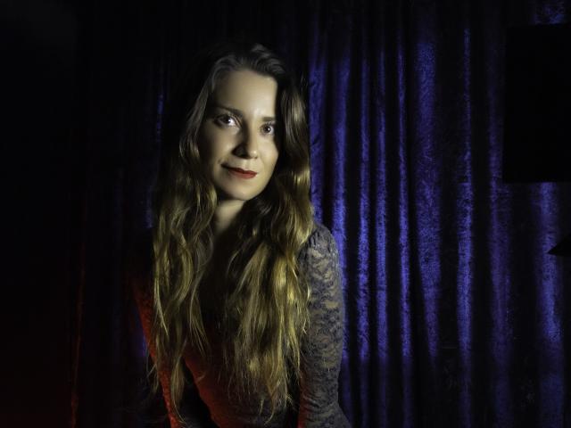 NatashaKute a de longs cheveux bruns ondulés, elle porte un haut en dentelle, avec un sourire discret, et se tient devant des rideaux violet foncé, éclairés doucement..