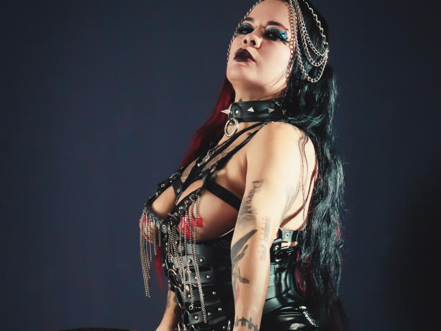 MorganaCrowly with black leather harness, chains, and red accents, dark lipstick, tattoos, and long black-red hair, wearing a black choker, looking up with a confident, sexy expression.