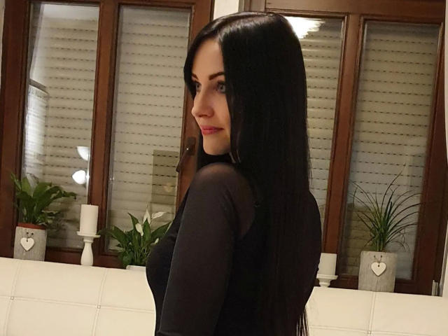 Joven, con una expresión de devoción, con cabello negro largo, ojos azules y labios rosados, vestida con una blusa negra transparente, mirando por encima del hombro, sonriendo ligeramente, con velas blancas y plantas en el fondo..