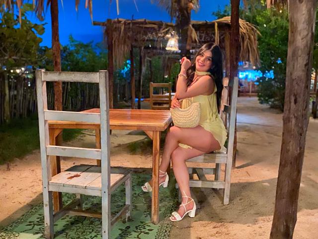 PaulinaValley, sitting on a wooden chair, wearing a yellow dress, white sandals, and a straw bag, with her ass facing the camera, in a beachside setting at night.