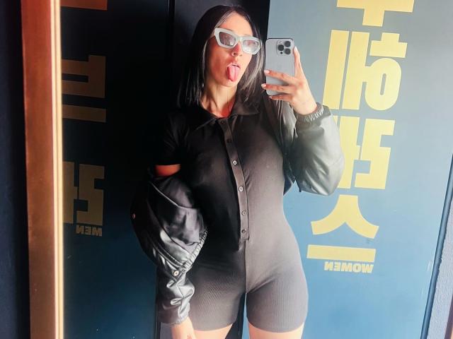 NicoColeman with black shirt and shorts, green jacket sleeves, white glasses, sticking tongue out, holding phone, mirror selfie, showing small tits and ass, dark hair, blue background.