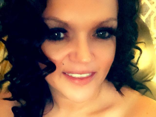 NaughtyWomanHot with curly black hair, green eyes, and a small beauty mark on her right cheek, smiling with white teeth. Her bare shoulders are visible.