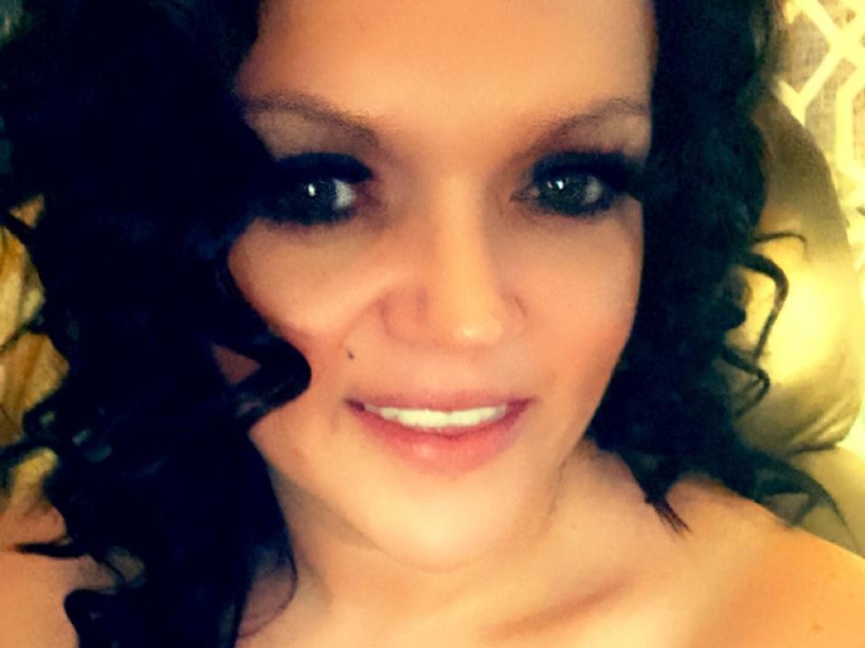 NaughtyWomanHot with curly black hair, green eyes, and a small beauty mark on her right cheek, smiling with white teeth. Her bare shoulders are visible.