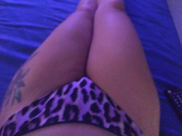 SeleneBright's legs are crossed, wearing leopard print panties, with a tattoo on her right thigh, lying on a blue bed, under purple lighting.