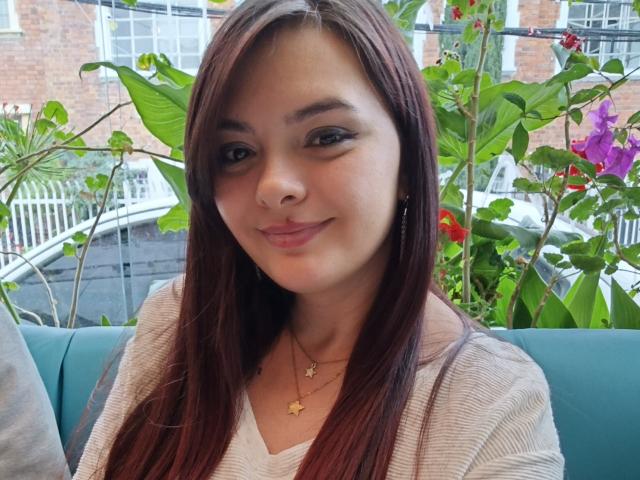 KatherinLatin heeft bruin haar, een schattig neusje en een subtiele glimlach. Ze draagt een witte top met een sterrenketting en zit op een turquoise bank met groene planten..