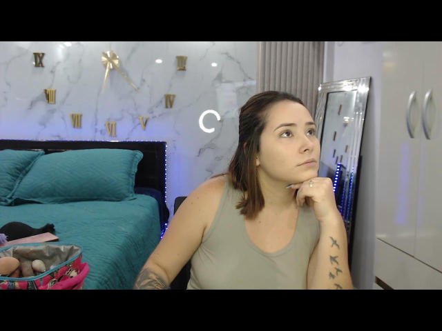 KarlaJonnes69 with brown hair, green tank top, tattoos on right arm, pensive expression, hand on chin, teal bed, mirror, and basket with toys in the bedroom.