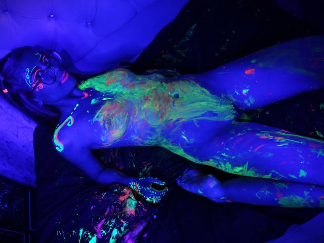 AbyColeman lies on a bed, covered in neon green, blue, and purple glow paint, with bright colors on her tits, pussy, and body, wearing glasses, in a blue-lit room.
