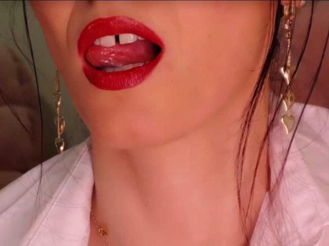 Het gezicht van BlueNightX, met rode lippenstift, haar tong uitgestoken en witte tanden. Ze draagt gouden oorbellen en een witte blouse, met een zichtbare gouden ketting..