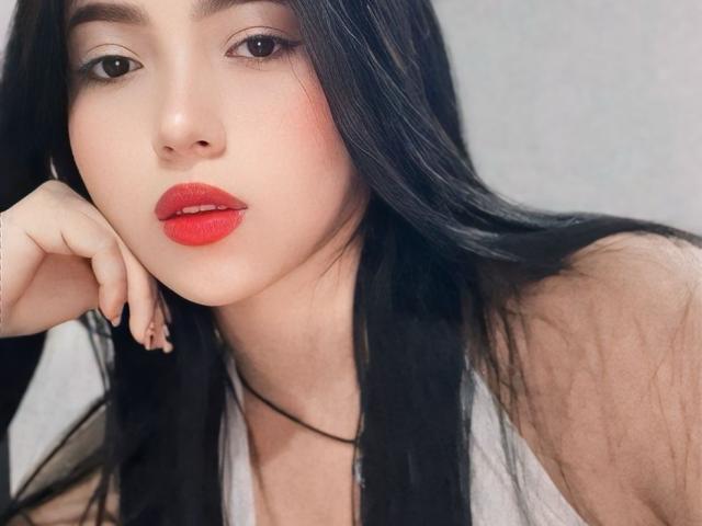 BellaDawson has long black hair, red lipstick, and a white tank top, resting her chin on her hand with a slight, sexy smile. Her nails are painted dark.