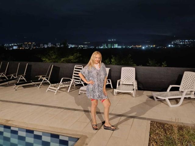 SunnyCleo with blonde hair, wearing a blue patterned dress, stands by a pool at night, surrounded by white lounge chairs, in sandals, under a dark sky.