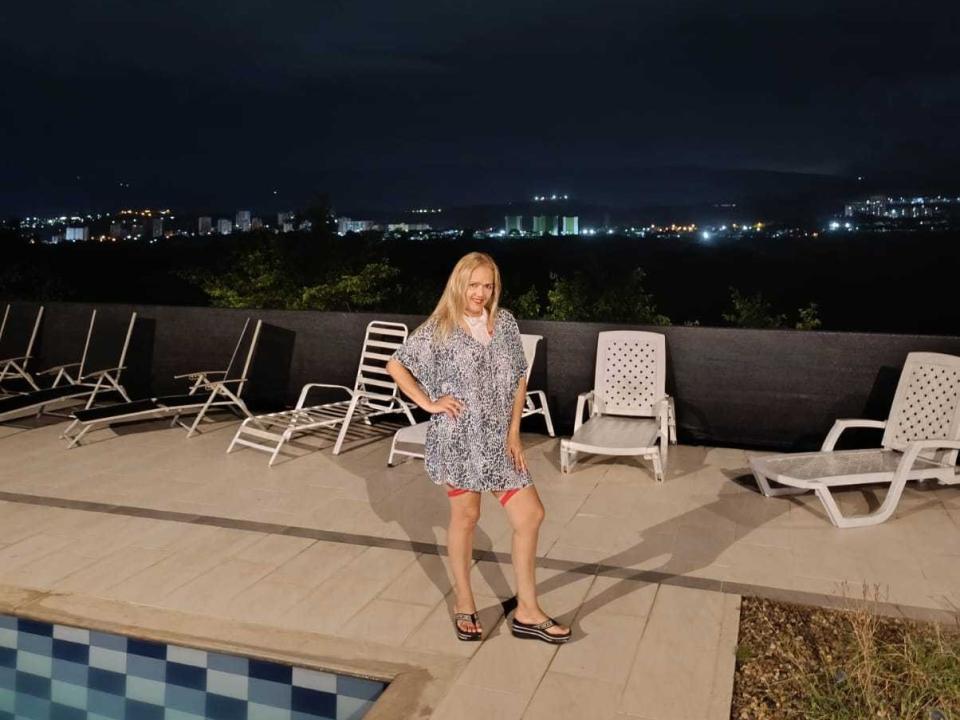 SunnyCleo with blonde hair, wearing a blue patterned dress, stands by a pool at night, surrounded by white lounge chairs, in sandals, under a dark sky.