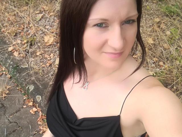 Sexyanny88-hot with pale skin, green eyes, and brown hair in a black one-shoulder top, wearing a heart necklace and hoop earrings, looking up with a slight smile.