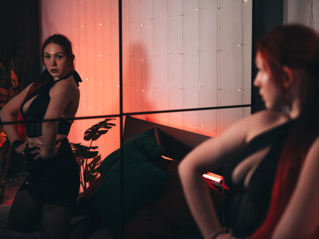 DoraXDora with red hair, black lingerie, and visible tits, standing and looking at her reflection in a mirror, hands on hips, in a dimly lit room with warm red lighting.