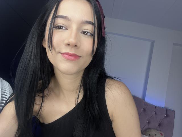 DeisyWest with long black hair, wearing a black tank top, pink lipstick, and a red hairband, smiling at the camera. Her tits are small, and she's cute.