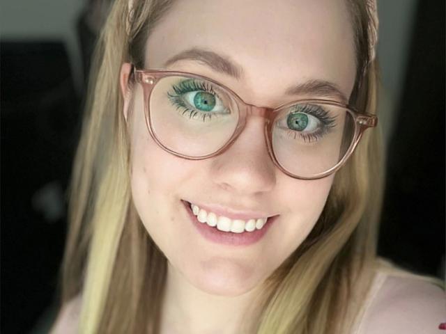 MiraMore, con occhi verdi, indossa occhiali marroni, sorride, ha i capelli biondi intrecciati, la pelle chiara e una camicia rosa, mostrando i suoi denti bianchi..