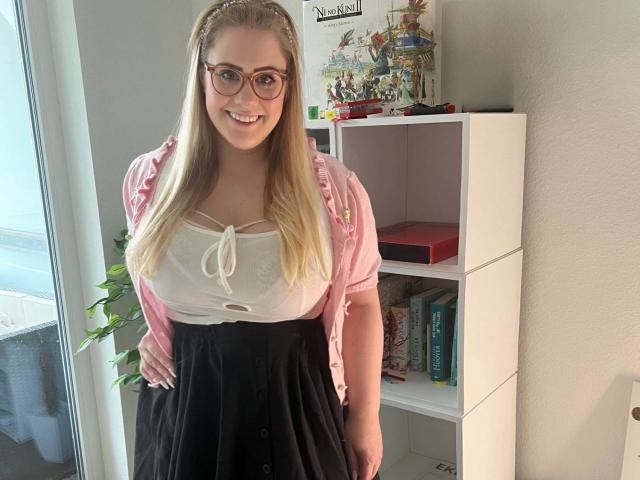 MiraMore-hot heeft blond haar, een bril, een roze vest, een witte, doorschijnende top en een zwarte rok, en ze glimlacht in een kamer met een witte plank en een plant. Borsten zijn zichtbaar onder de top..
