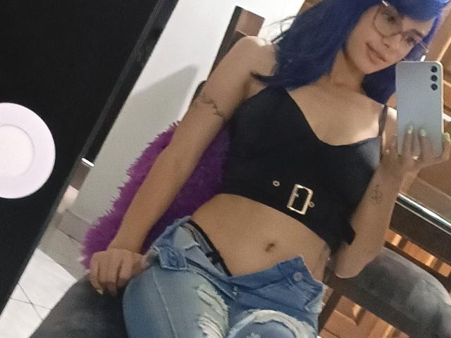 AnaisHilton with blue hair, glasses, black crop top, belt, and ripped jeans, showing her flat stomach and black underwear, taking a mirror selfie.