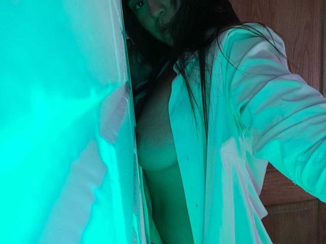SharongoldOne with long dark hair, wearing an open white shirt, exposing her tits, standing in a green-lit room, partially visible face, right arm raised, wooden door in background.