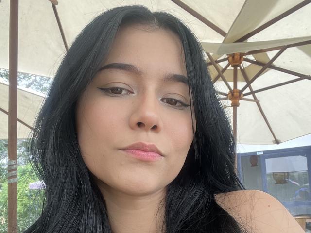 DeisyWest's face, with black hair, brown eyes, and pink lips, showing a slight pout. Her eyeliner is winged, and she's wearing a black top. Under a beige umbrella.