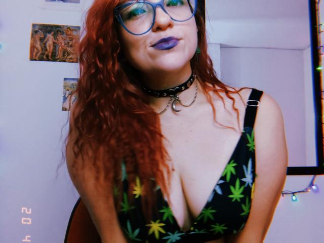 LunaRausch with red curly hair, blue glasses, purple lipstick, black choker, chain, deep cleavage in black dress with green marijuana leaves, smiling. Boobs visible.