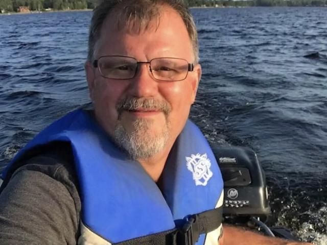 middle-aged dude with glasses, gray beard, blue life jacket, smiling at camera, sitting in boat, water motor in background, holding camera, slightly tan, wearing gray shirt.